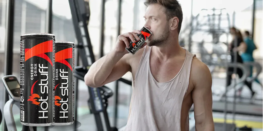 A fit man who drinks Hot Stuff Energy Drink in a gym, with workout equipment and large windows in the background.